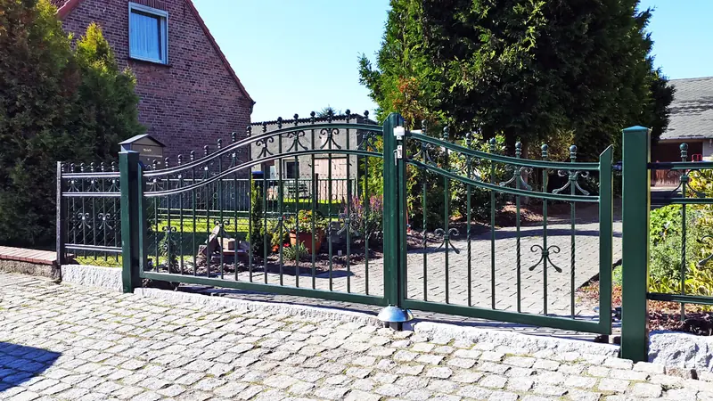 Dunkelgrünes schmiedeeisernes Hoftor mit Bogen und Zierspitzen auf gepflastertem Hof